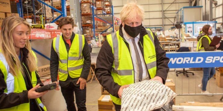 ritský premiér Boris Johnson s ochranným rúškom (vpredu) drží balík počas jeho návštevy v sklade športového obchodu Next World Sports v anglickom meste Wrexham 26. apríla 2021.