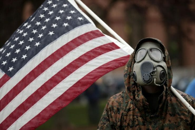 Demonštrant s plynovou maskou na tvári drží americkú vlajku na zhromaždení proti nariadeniu štátu Michigan, ktorým žiada občanov, aby zostali doma počas koronavírusovej pandémie v meste Lansing vo štvrtok 14. mája 2020.